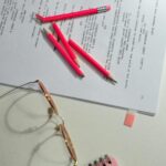 Still life of a workspace with pink pencils, papers, and glasses on a desk.